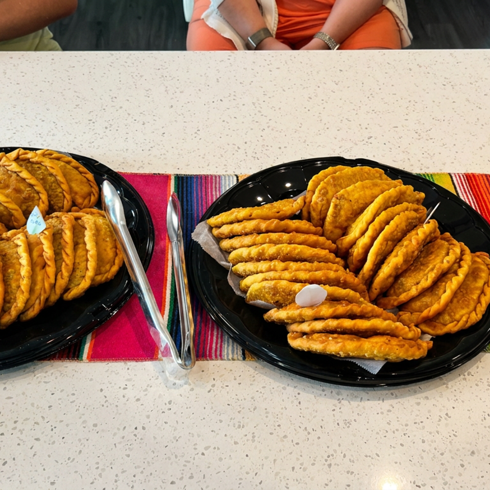 Two black platters hold assorted golden fried empanadas on a colorful striped tablecloth, with dipping sauces and diners in the background.