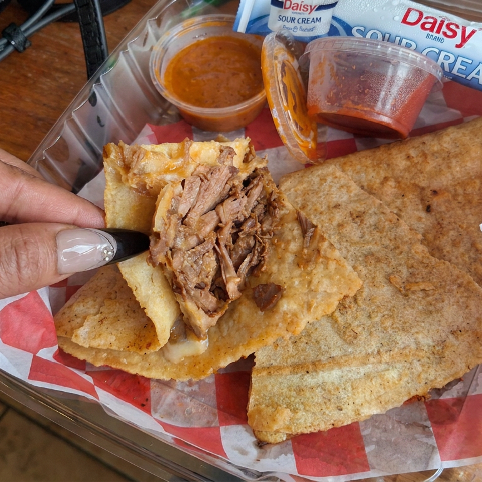 Hand holds a crispy folded quesadilla filled with shredded meat, served with red and orange sauces, sour cream, and receipt.